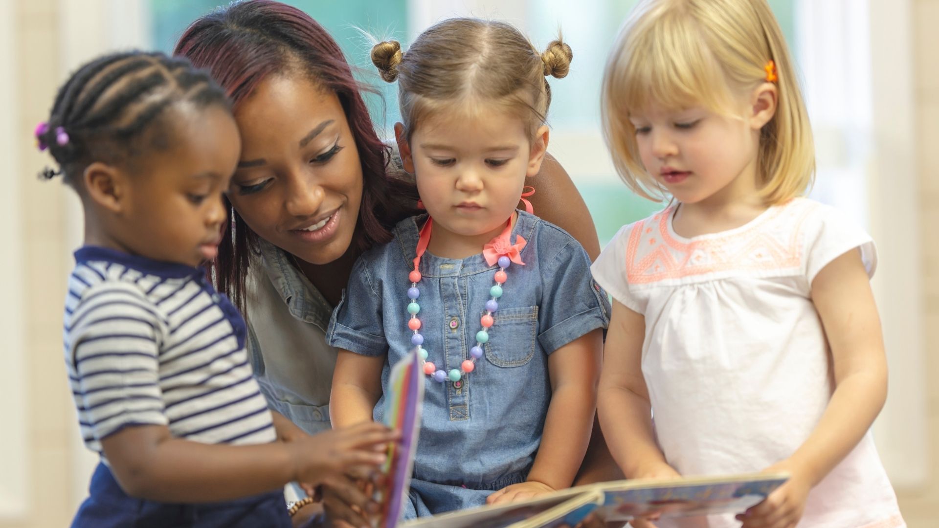 A female teacher with three preschool ages girls reading a book.