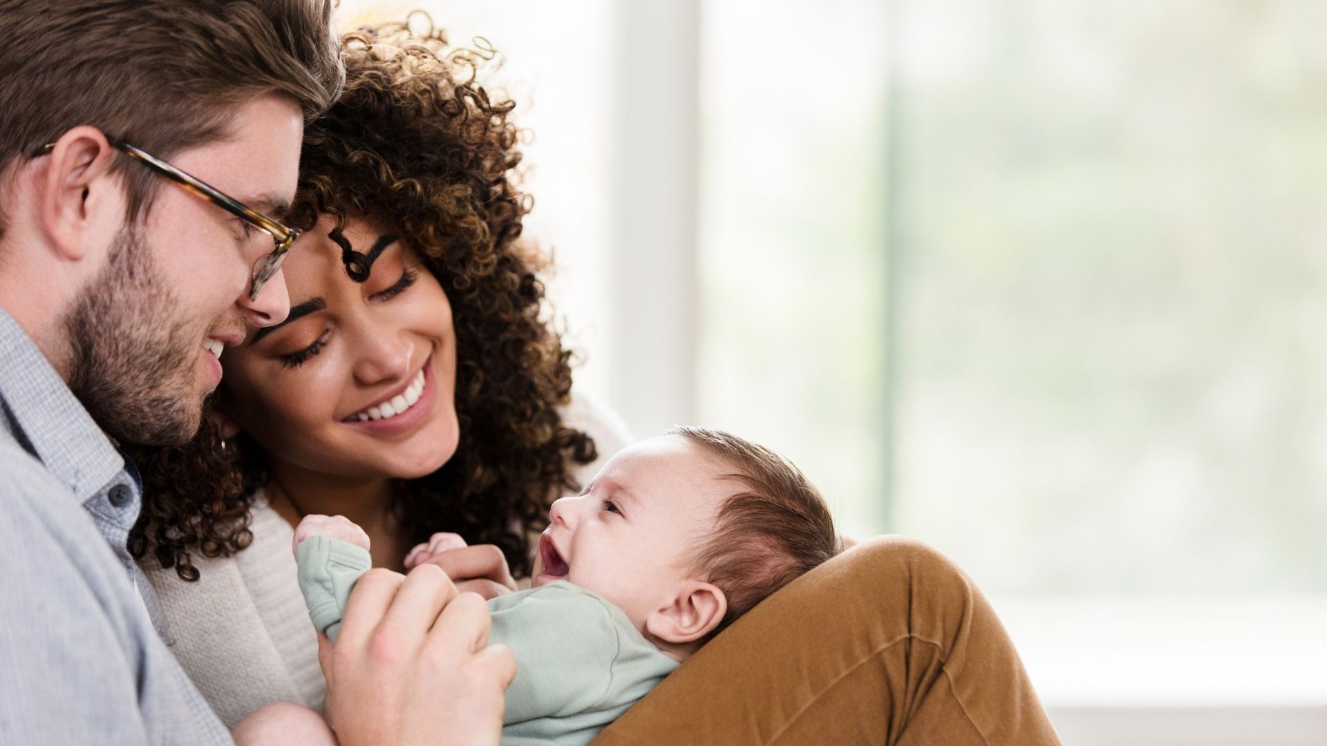 A father with short brown hair and glasses, and a mother with dark brown curly hair both looking down and smiling at a smiling baby who is looking up at them. The baby is on the father’s lap with the knees propped up.