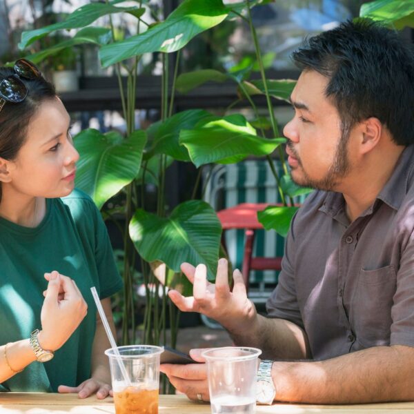 A man and a woman sitting at a coffee shop with plants in the background. The woman has sunglasses in her hair and is wearing a green shirt. And the male is wearing a grey shirt.