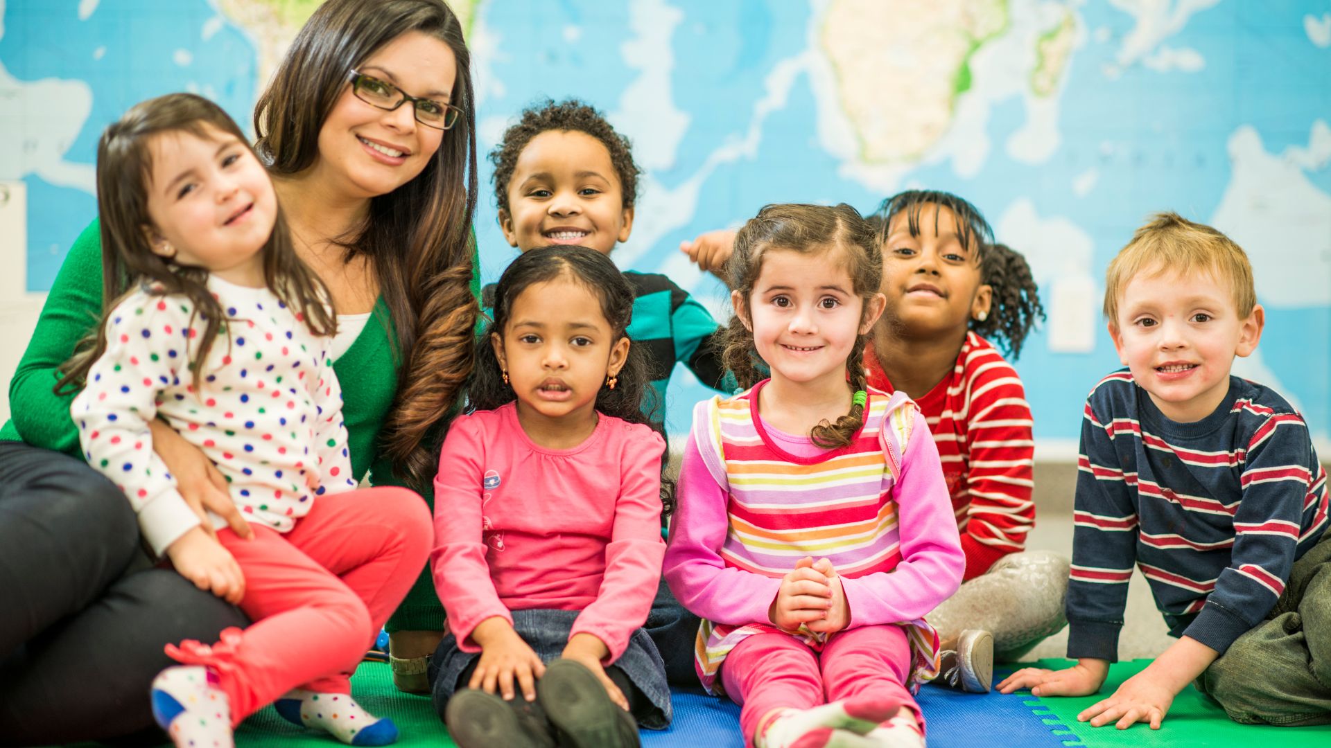A female teacher with glasses sitting in her classroom surrounded by six children smiling at the camera with a map of the world as their background.