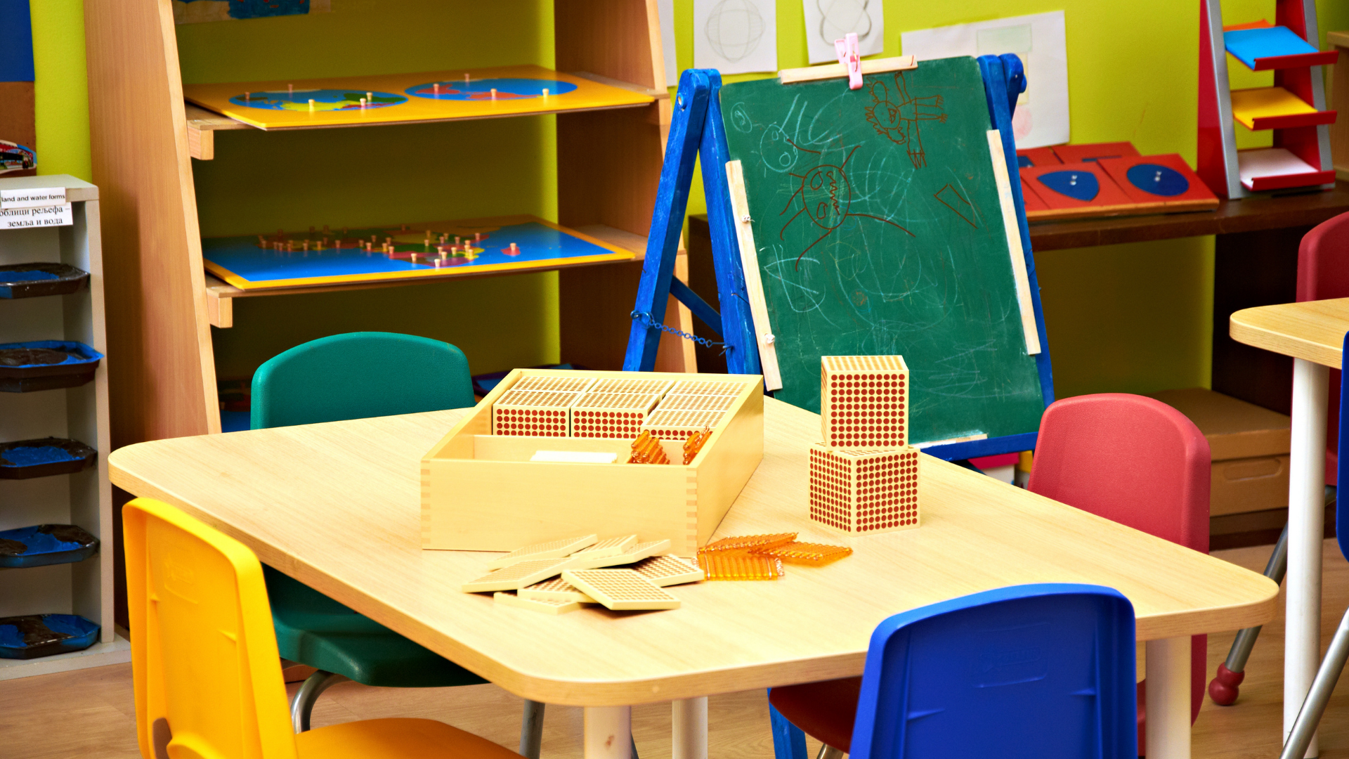 An image of a close up on a small table with four colorful small chairs surrounding it and on top of the table there are blocks. There’s also an easel with green chalkboard for children to play with.