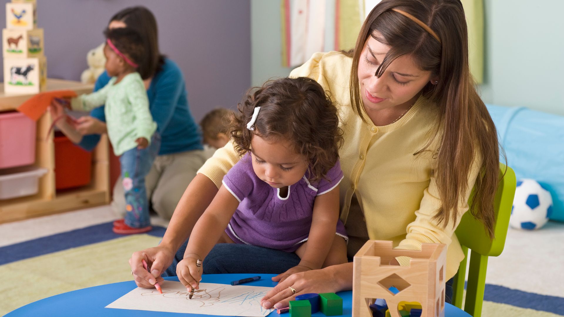 An image of a female teacher and young child who are drawing together at the desk in a classroom. Another female teacher and two young children are seen in the background.