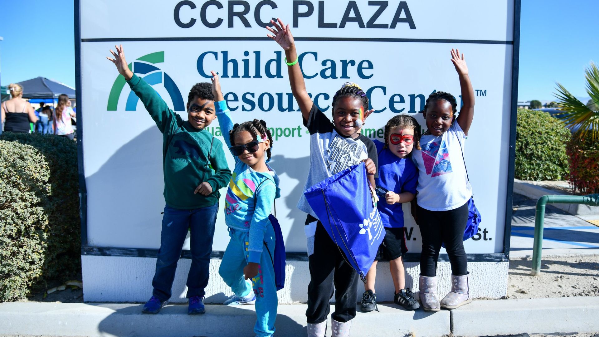 Five children raising their hands in the air and smiling in front of the Child Care Resource Center sign located outside of our CCRC Victorville Office
