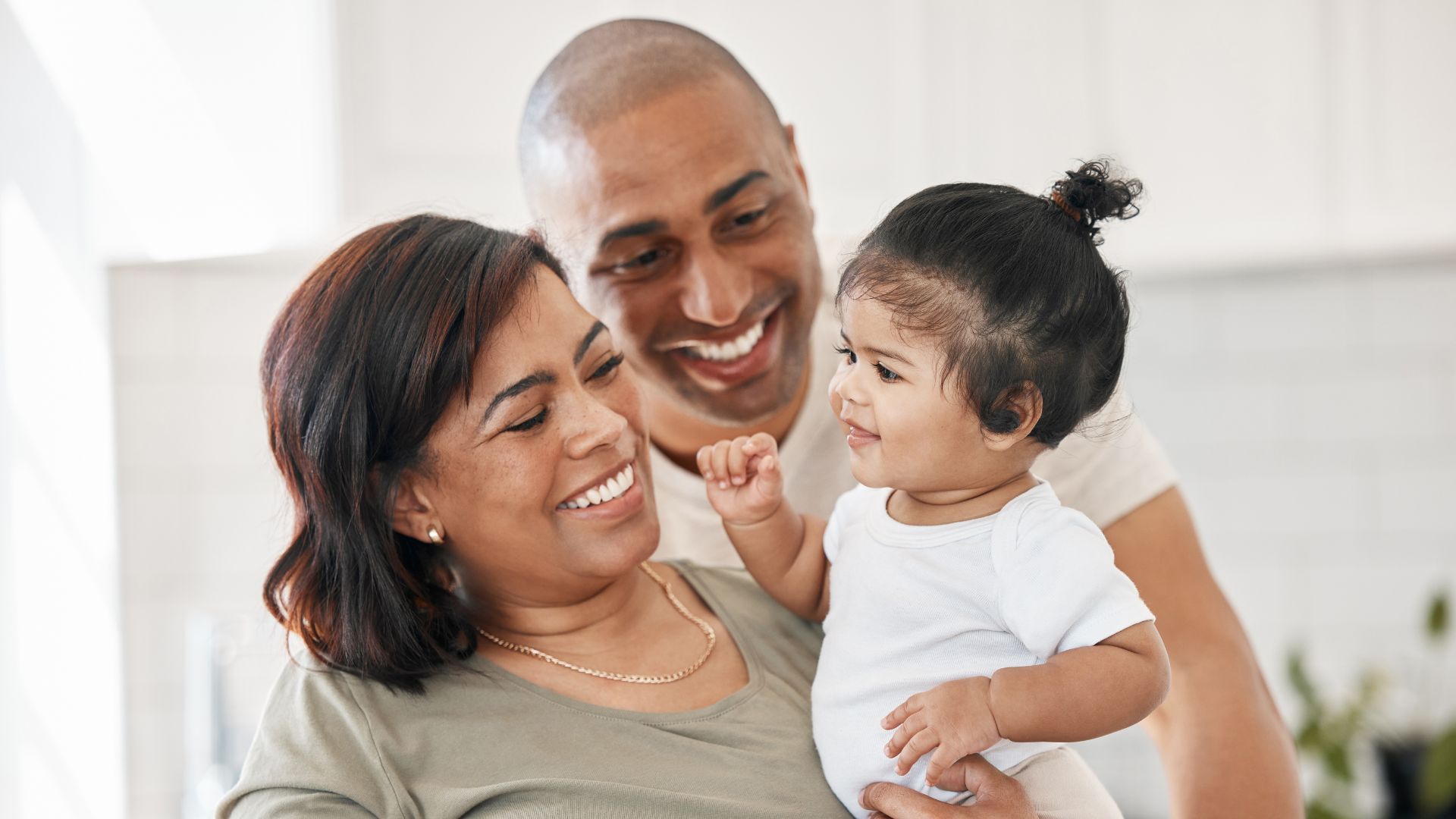 A mother smiles at her infant daughter that she's holding. Dad is smiling at the infant daughter as well.