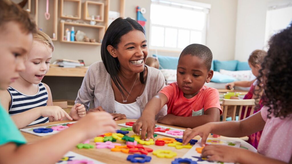 Female teacher surrounded by two children on either side of her at a kids table while the children are playing with colorful gears.