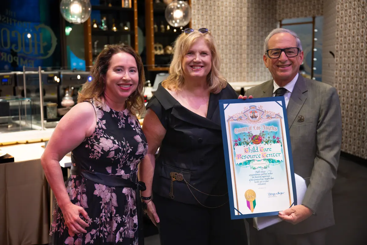 Monica Banken, Donna Sneeringer, and Dr. Michael Olenick smiling inside the The Toothsome Chocolate Emporium & Savory Feast Kitchen with Dr. Olenick holding a commemorative County of Los Angeles, Child Care Resource Center scroll