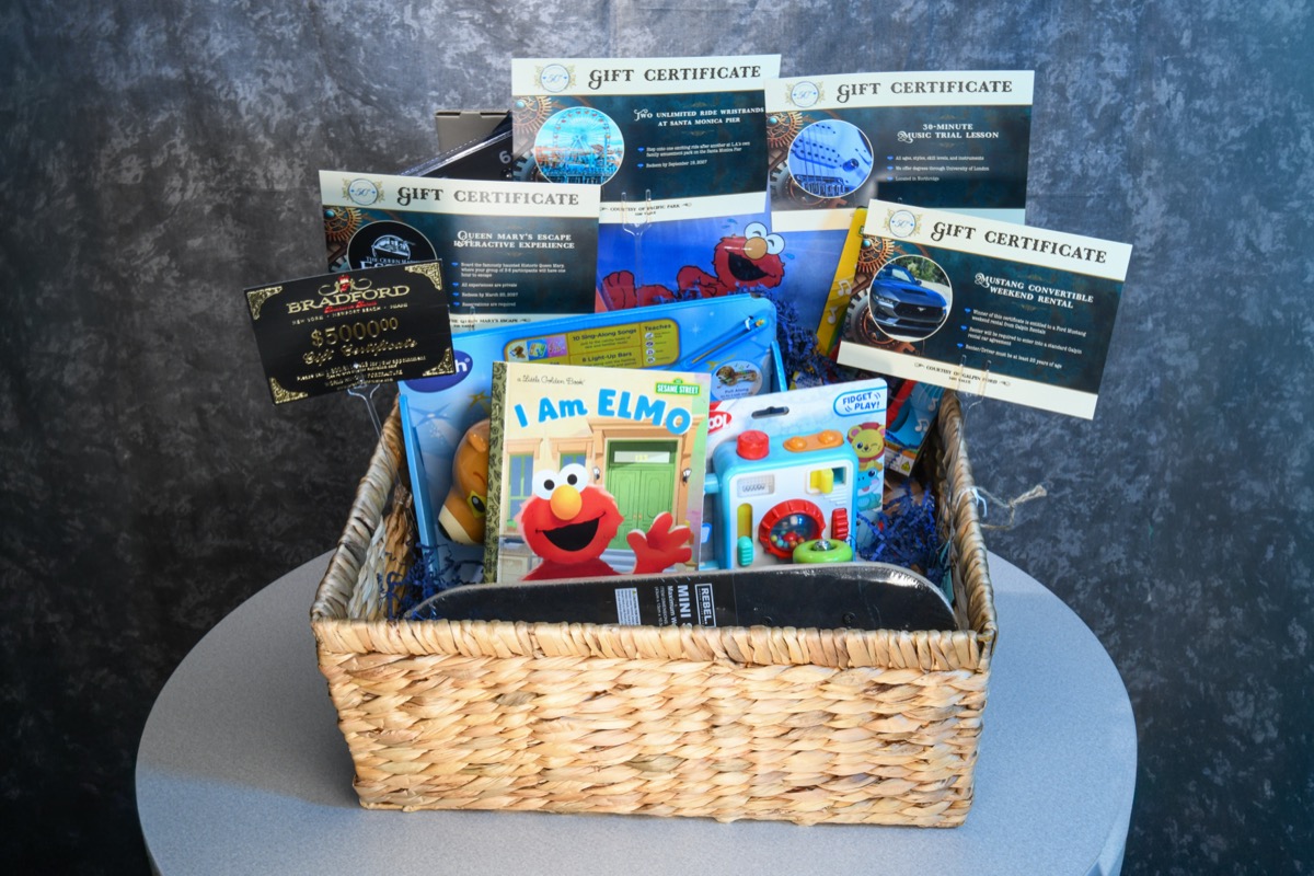 A wicker basket filled with various prizes, including gift certificates for experiences and a children's book titled 'I Am Elmo', sits on a gray table against a dark textured background.
