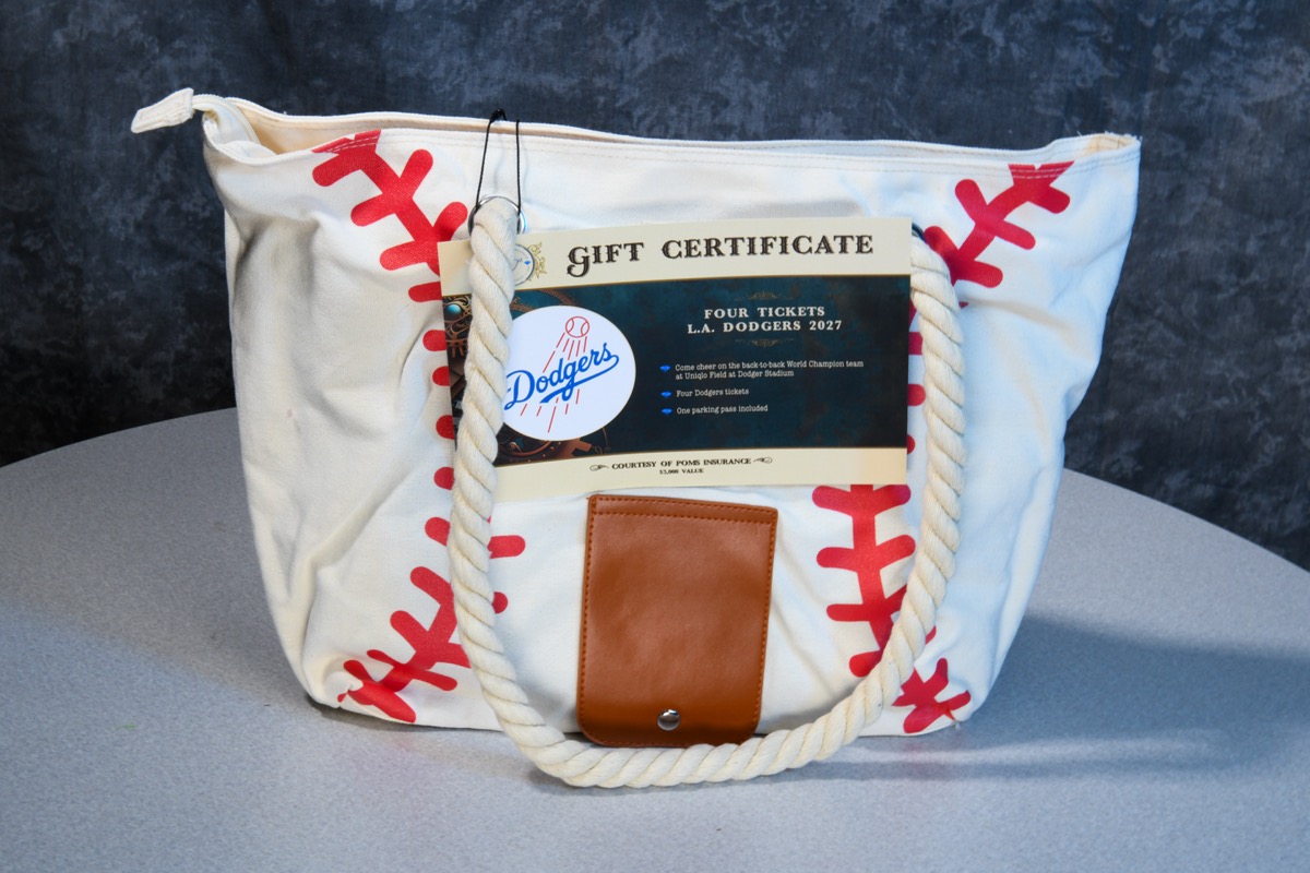 A white canvas tote bag with red stitching resembling a baseball is displayed on a gray surface, featuring a gift certificate for four L.A. Dodgers 2027 tickets. The bag has thick rope handles and a brown leather patch, set against a dark textured background.