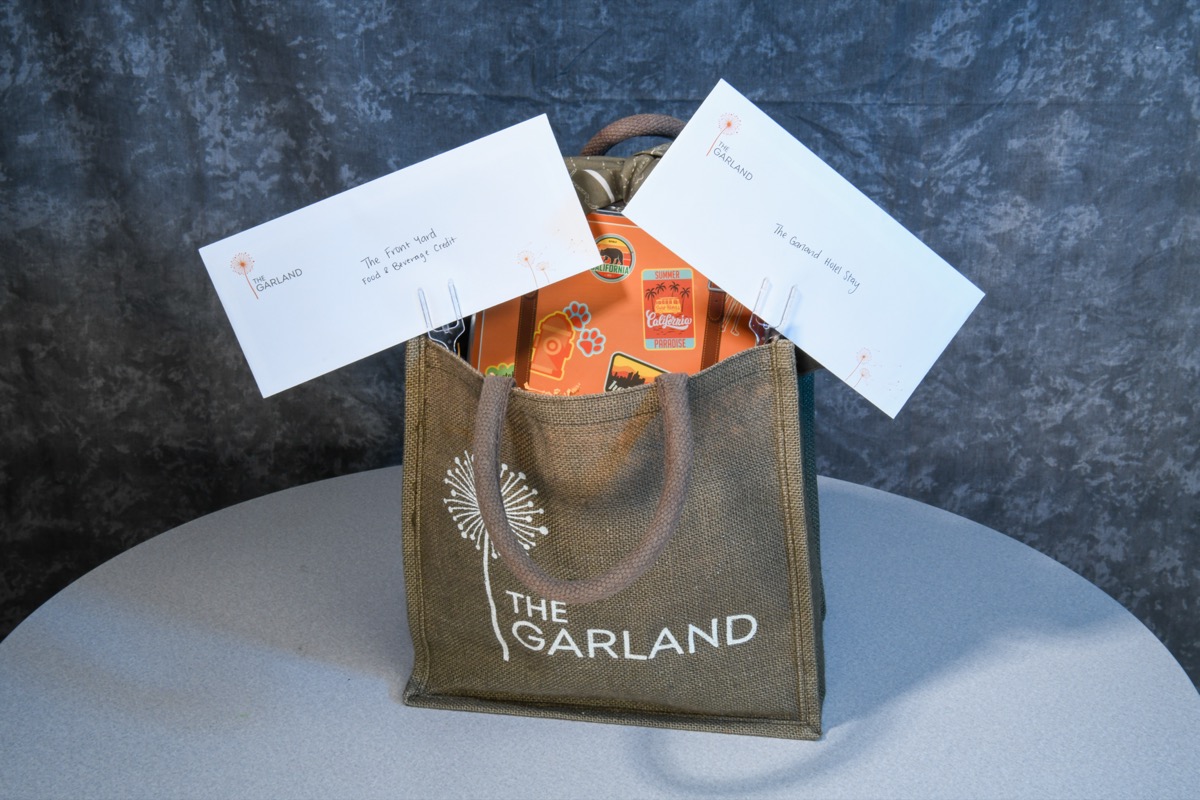 A brown tote bag with "The Garland" printed on it sits on a gray table, filled with items including two white envelopes and an orange booklet.