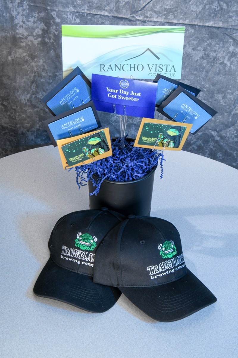 A black bucket filled with blue shredded paper and multiple gift cards, including some from Antelope Valley Mall, sits on a white table. Two black baseball caps featuring the Transplants Brewing Company logo are placed in front of it, with a Rancho Vista Golf Club sign visible behind.