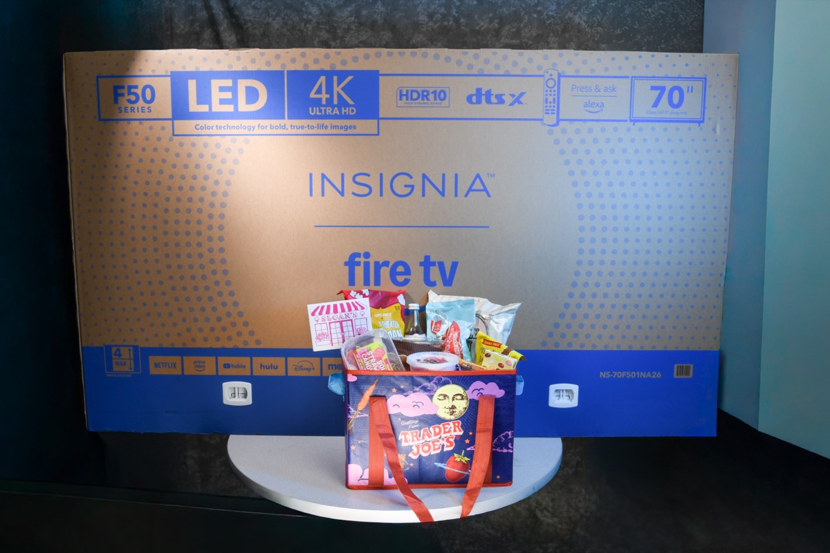 A large box for an Insignia 70-inch LED 4K Ultra HD Fire TV sits on a white pedestal, with a Trader Joe's branded tote bag filled with snacks placed in front of it.