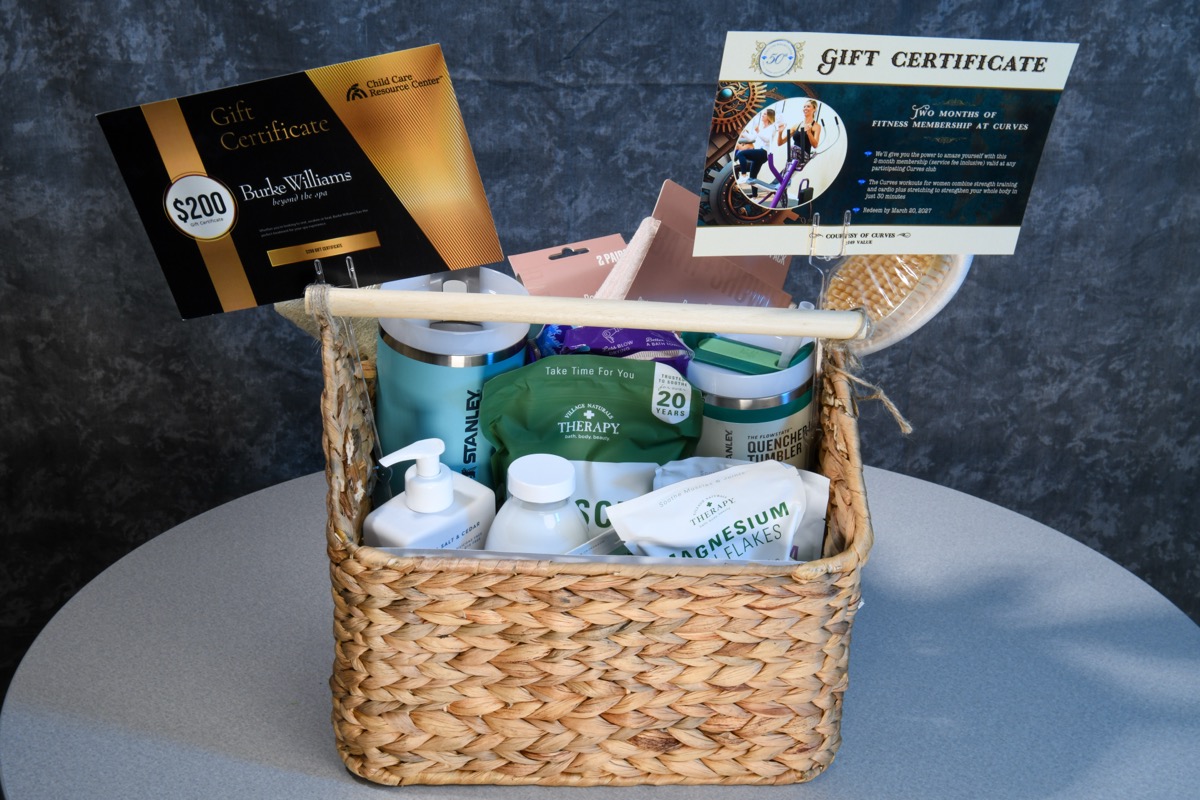 A woven basket filled with various spa and wellness items, including gift certificates for Burke Williams and Curves, sits on a gray table against a dark textured wall. The basket contains products like magnesium flakes, therapy bottles, and branded containers.