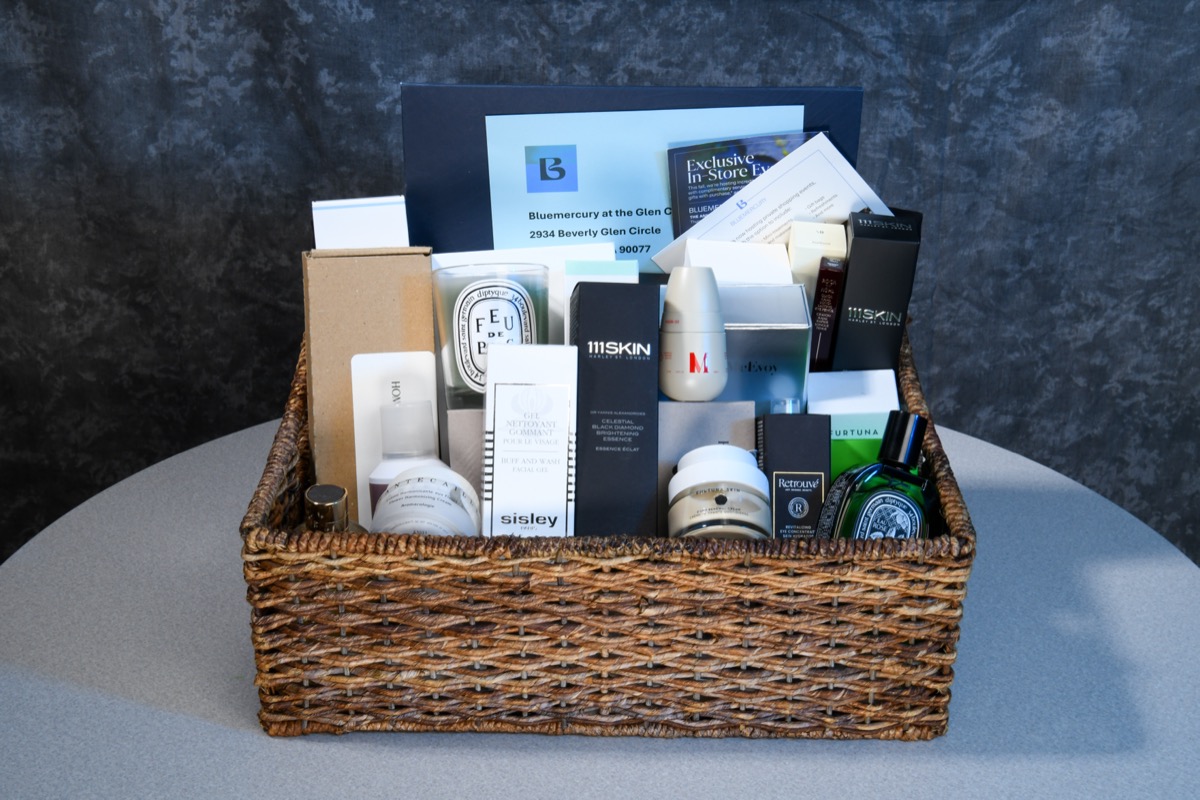 A wicker basket filled with various luxury beauty and skincare products, including items from brands like Sisley, 111SKIN, and Diptyque, sits on a gray table against a dark textured wall.